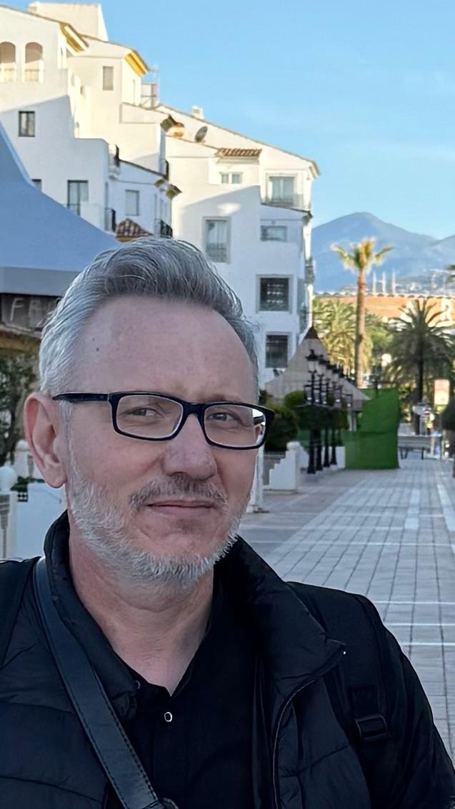 Portret mężczyzny w okularach na tle białych budynków z palmami i górami w oddali, w słoneczny dzień. Ujęcie z dołu, widoczny fragment chodnika.