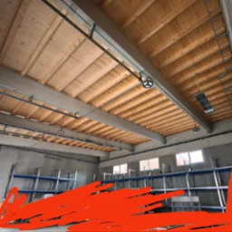 Wnętrze hali magazynowej z drewnianym stropem, oświetleniem przemysłowym i metalowymi regałami. Widoczne instalacje pod sufitem i drabina na ścianie.