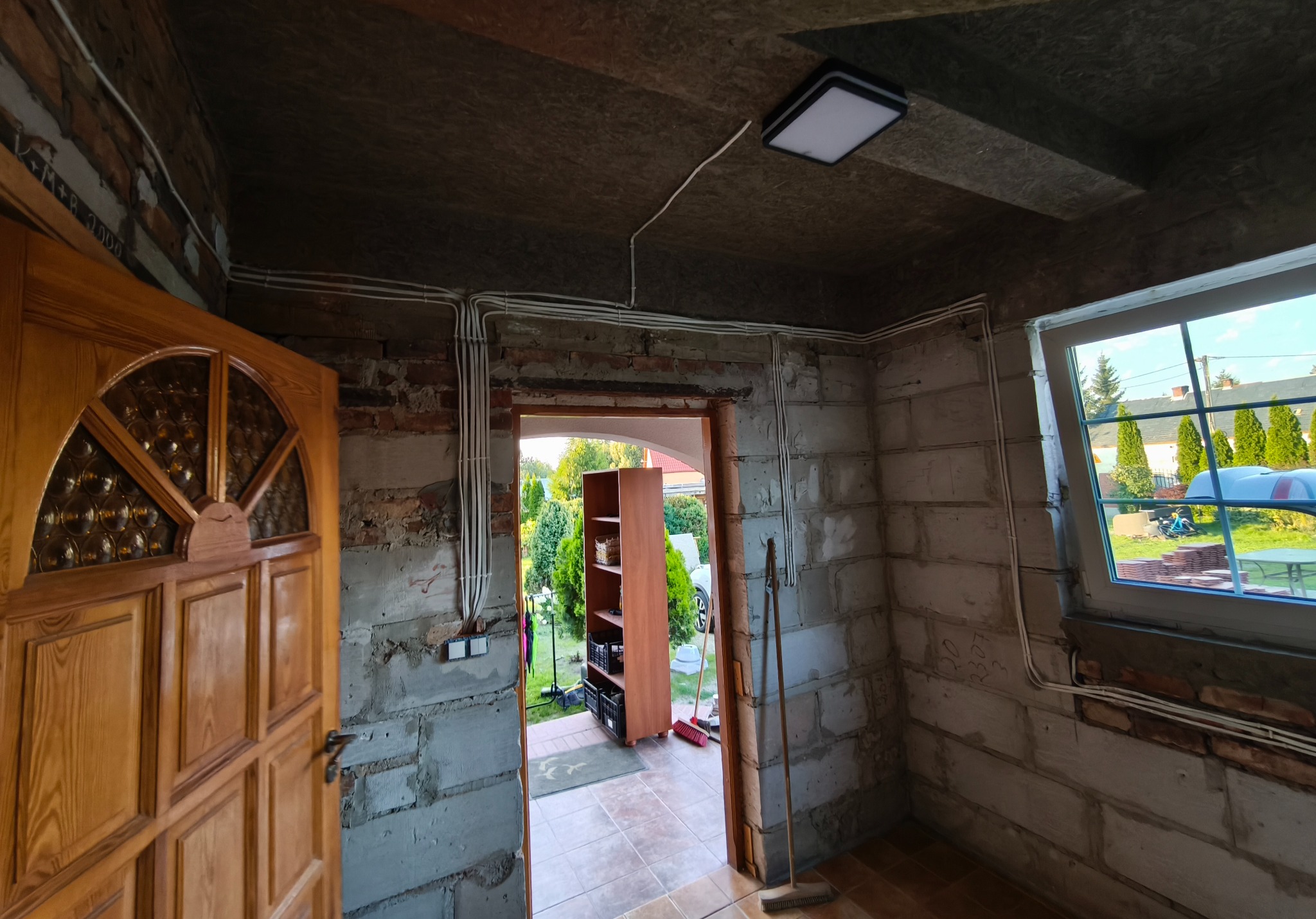 Natynkowa instalacja elektryczna w surowym wnętrzu przed tynkowaniem. Widoczne przewody, puszki i włączniki. Okno z widokiem na zieleń. Drzwi wejściowe z szybą.