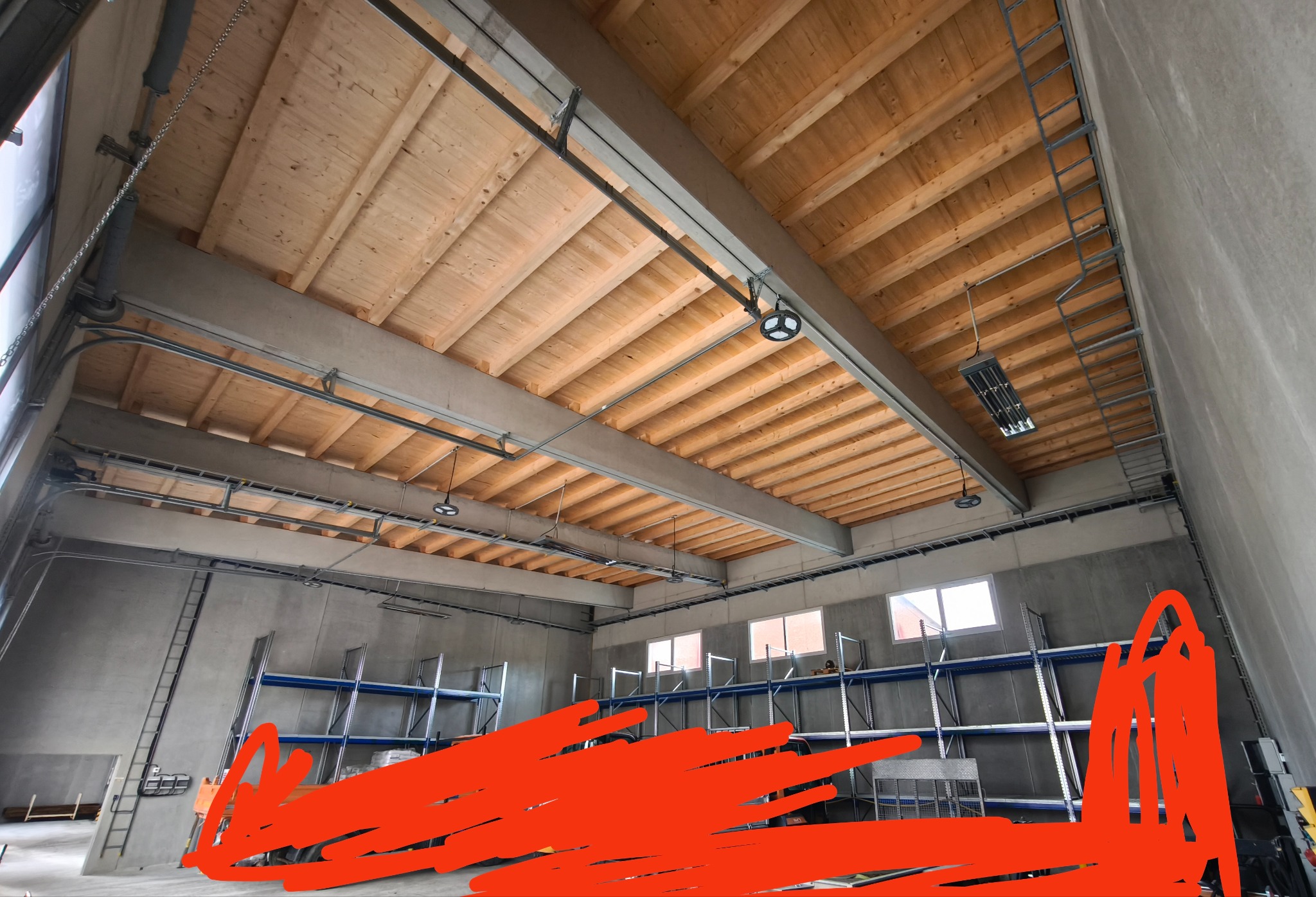 Wnętrze hali magazynowej z drewnianym stropem, oświetleniem przemysłowym i metalowymi regałami. Widoczne instalacje pod sufitem i drabina na ścianie.