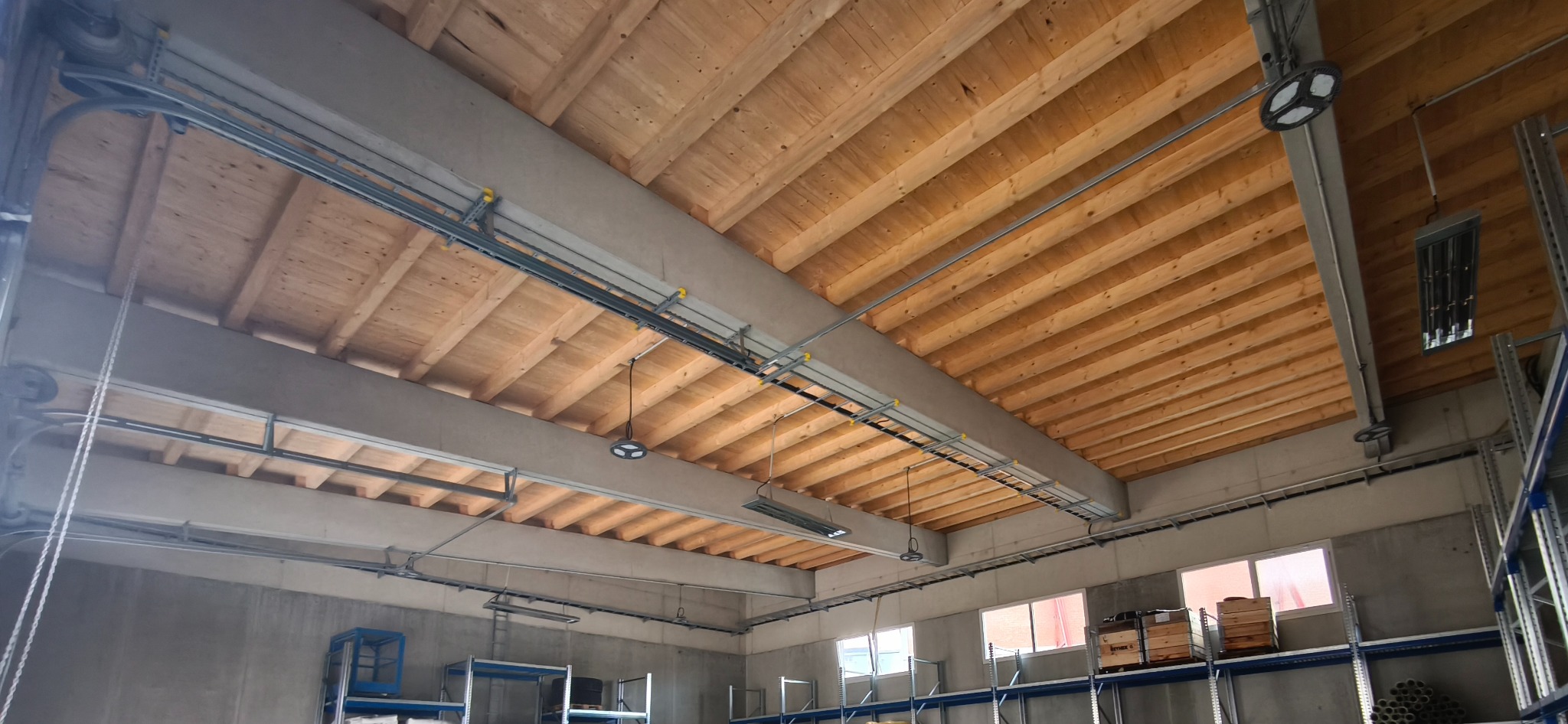 Instalacje elektryczne pod drewnianym stropem w hali magazynowej. Widoczne koryta kablowe, przewody i lampy LED. Ujęcie z dołu, szeroki kadr.