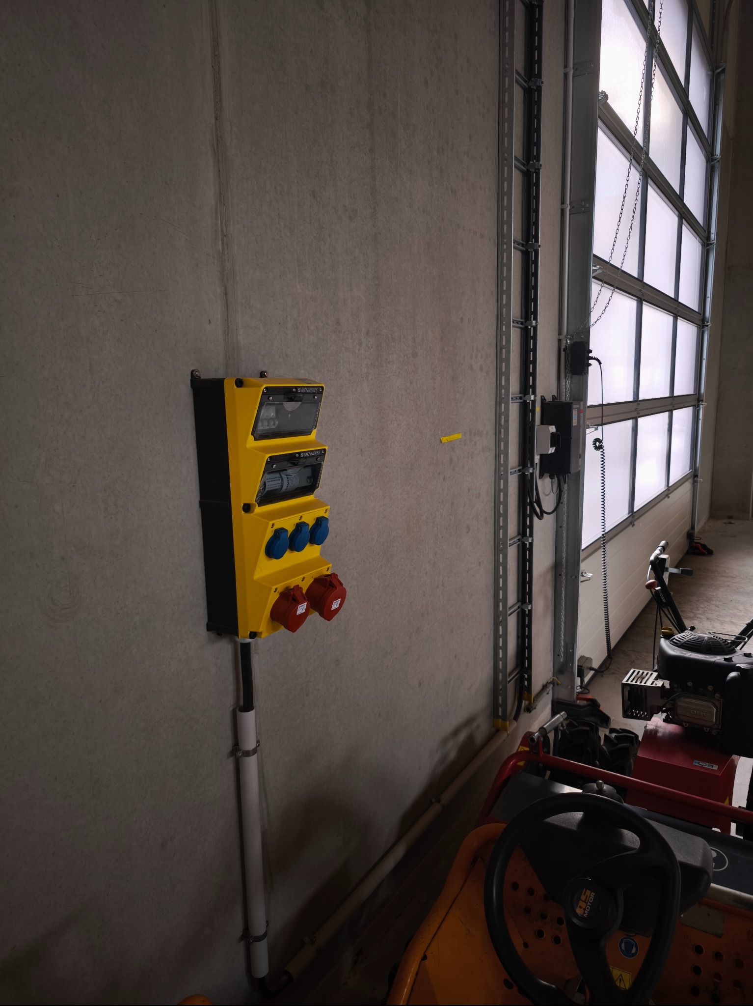Żółta, natynkowa rozdzielnica elektryczna z gniazdami siłowymi na szarej ścianie w hali, obok bramy segmentowej i sprzętu ogrodniczego.