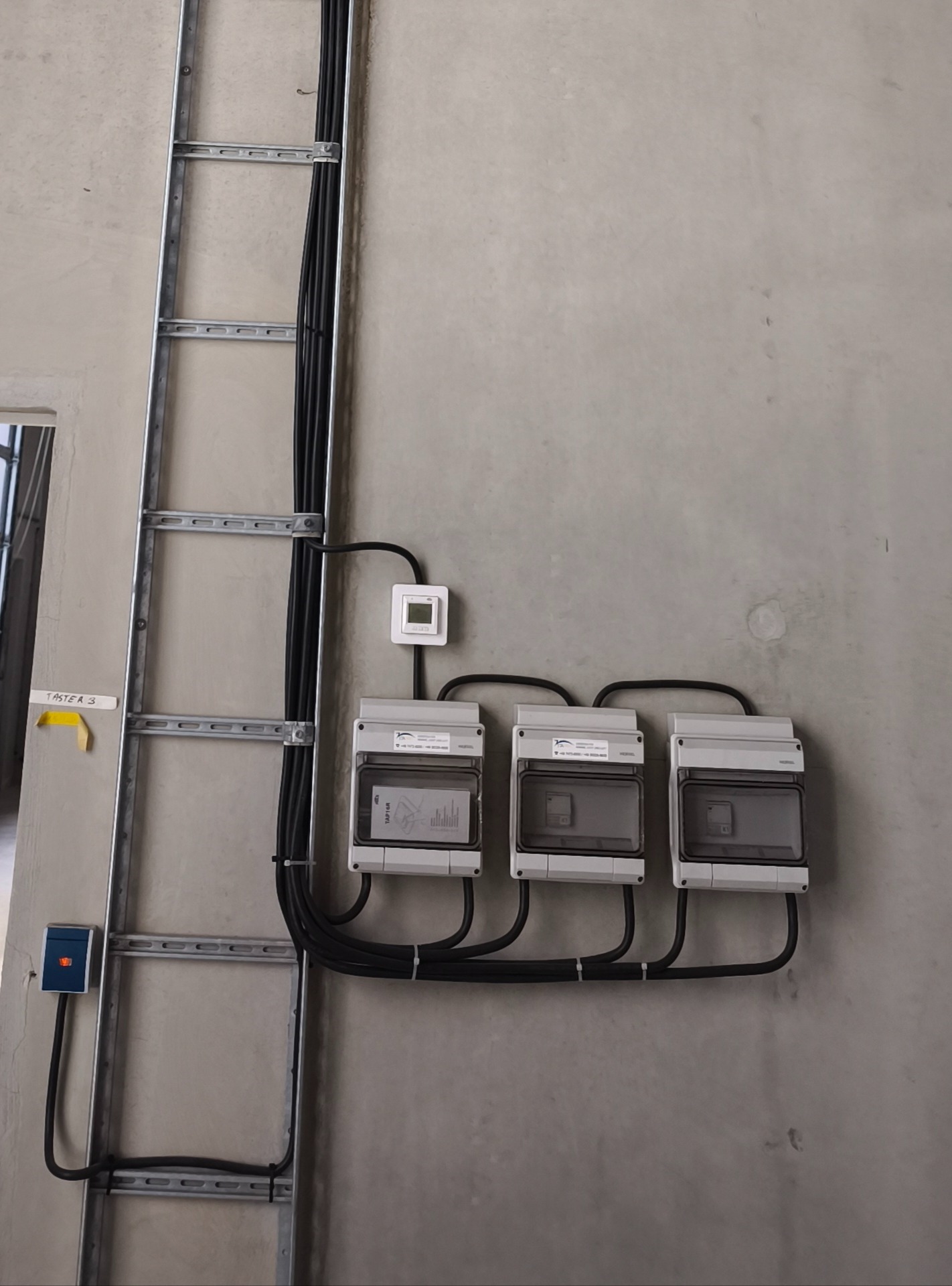 Szara ściana z trzema szarymi skrzynkami elektrycznymi, czarnymi kablami i metalową drabinką kablową. Widoczny mały, biały termostat.