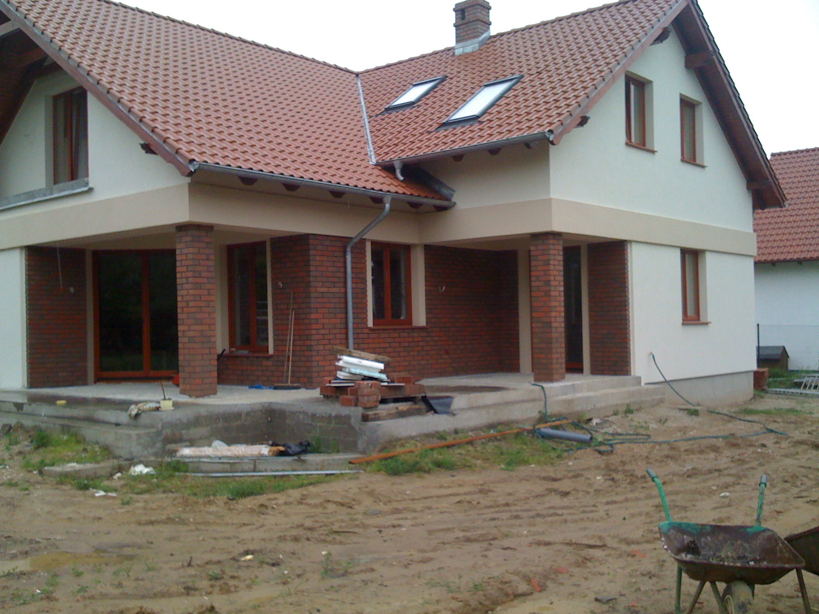 Nowy dom w trakcie wykończenia z czerwoną dachówką, ceglanymi kolumnami werandy i widocznymi materiałami budowlanymi na zewnątrz.