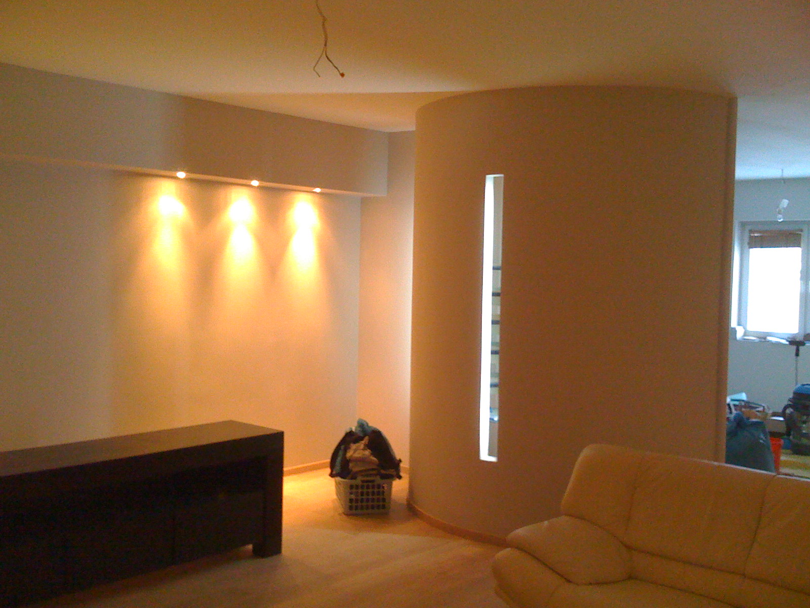 Wnętrze pokoju po remoncie z oświetleniem punktowym na ścianie, zaokrąglonym narożnikiem z pionowym oknem, częściowo widoczną żółtą sofą i koszem z praniem w rogu