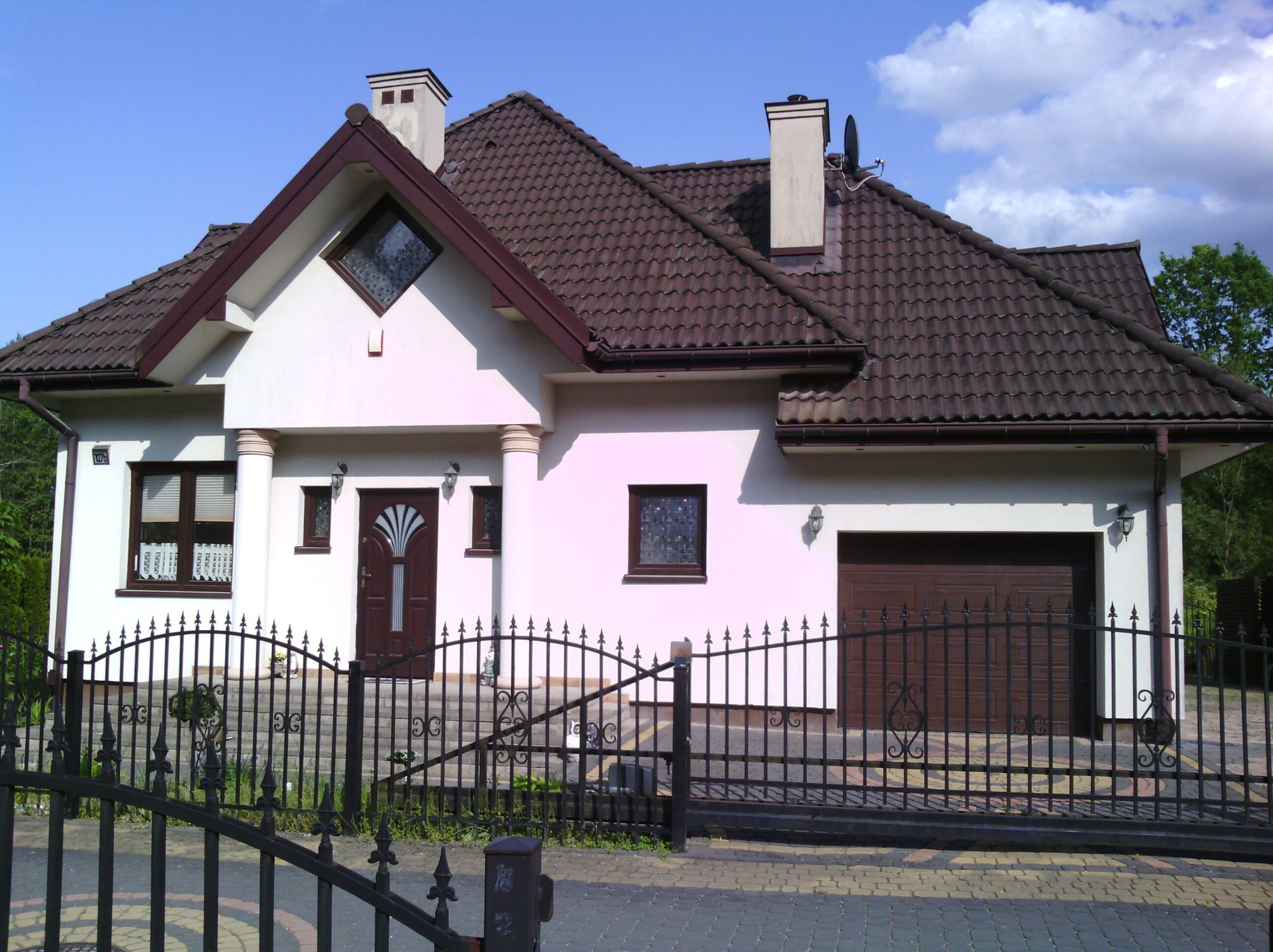 Dom jednorodzinny z brązowym dachem i jasną elewacją w Górznie, widziany zza czarnego, ozdobnego ogrodzenia. Widoczne detale architektoniczne i błękitne niebo.