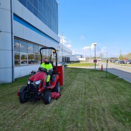 Service Creative Green Transport Marcel Kopaniszyn - Czerwona kosiarka traktorek prowadzona przez operatora w odblaskowej kamizelce na tle nowoczesnego biurowca i parkingu. Widoczny efekt koszenia.