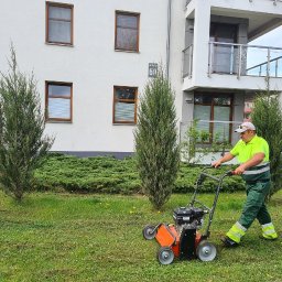 Service Creative Green Transport Marcel Kopaniszyn - Mężczyzna w odblaskowej kamizelce kosiarką spalinową pielęgnuje trawnik przed nowoczesnym budynkiem z balkonem i ozdobnymi iglakami.
