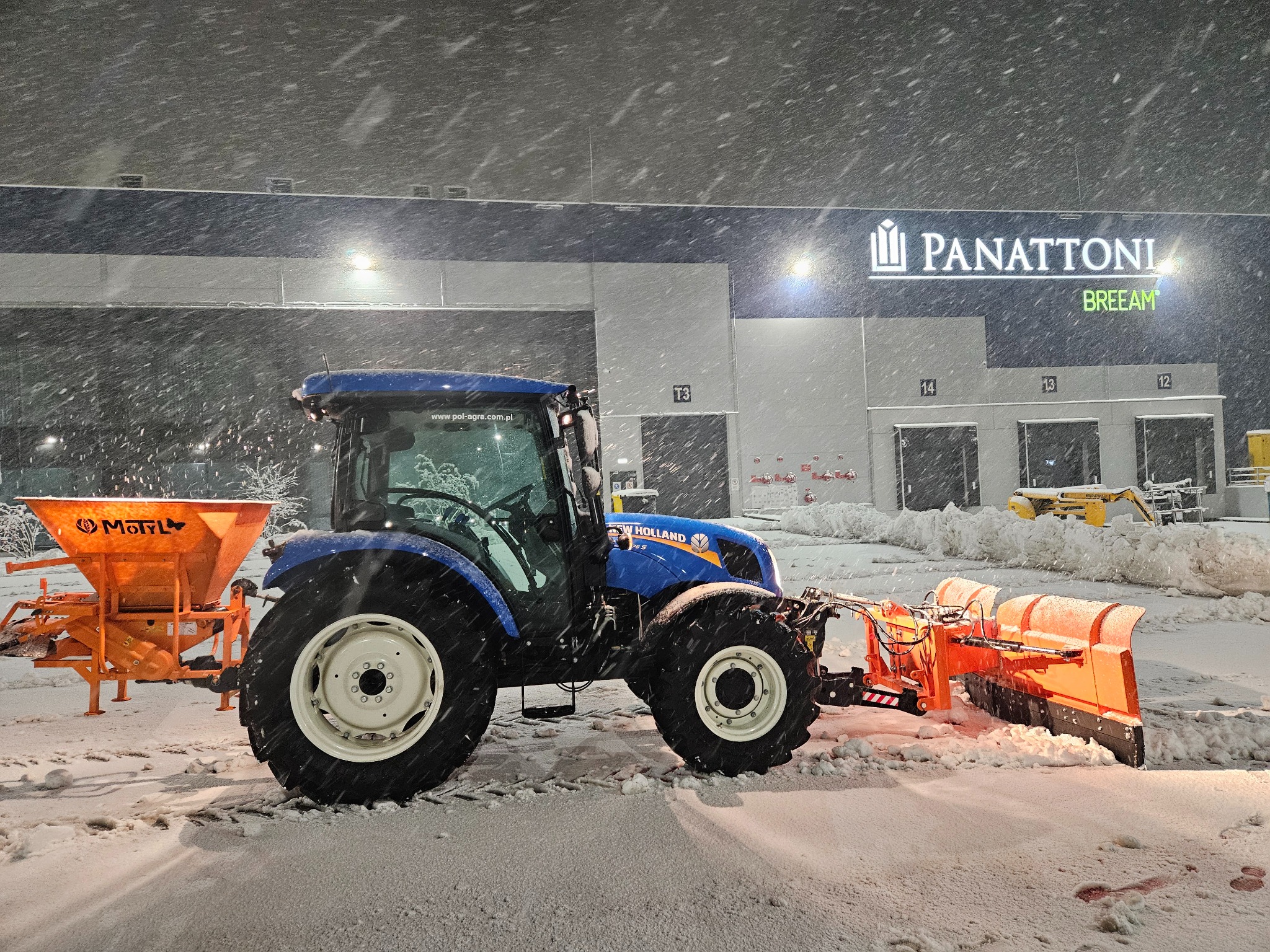 Niebieski traktor New Holland z pomarańczowym pługiem i posypywarką soli odśnieża teren przed halą Panattoni w trakcie intensywnych opadów śniegu.