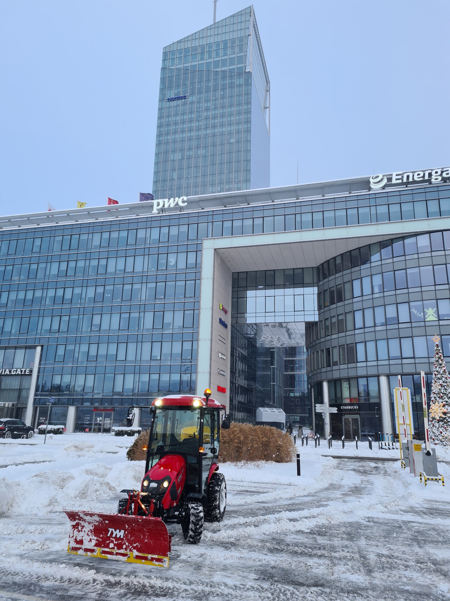 Czerwony traktor TYM z pługiem odśnieża plac przed nowoczesnym biurowcem z logo PwC i Energii, w tle choinka i wysoki wieżowiec. Zimowy dzień.