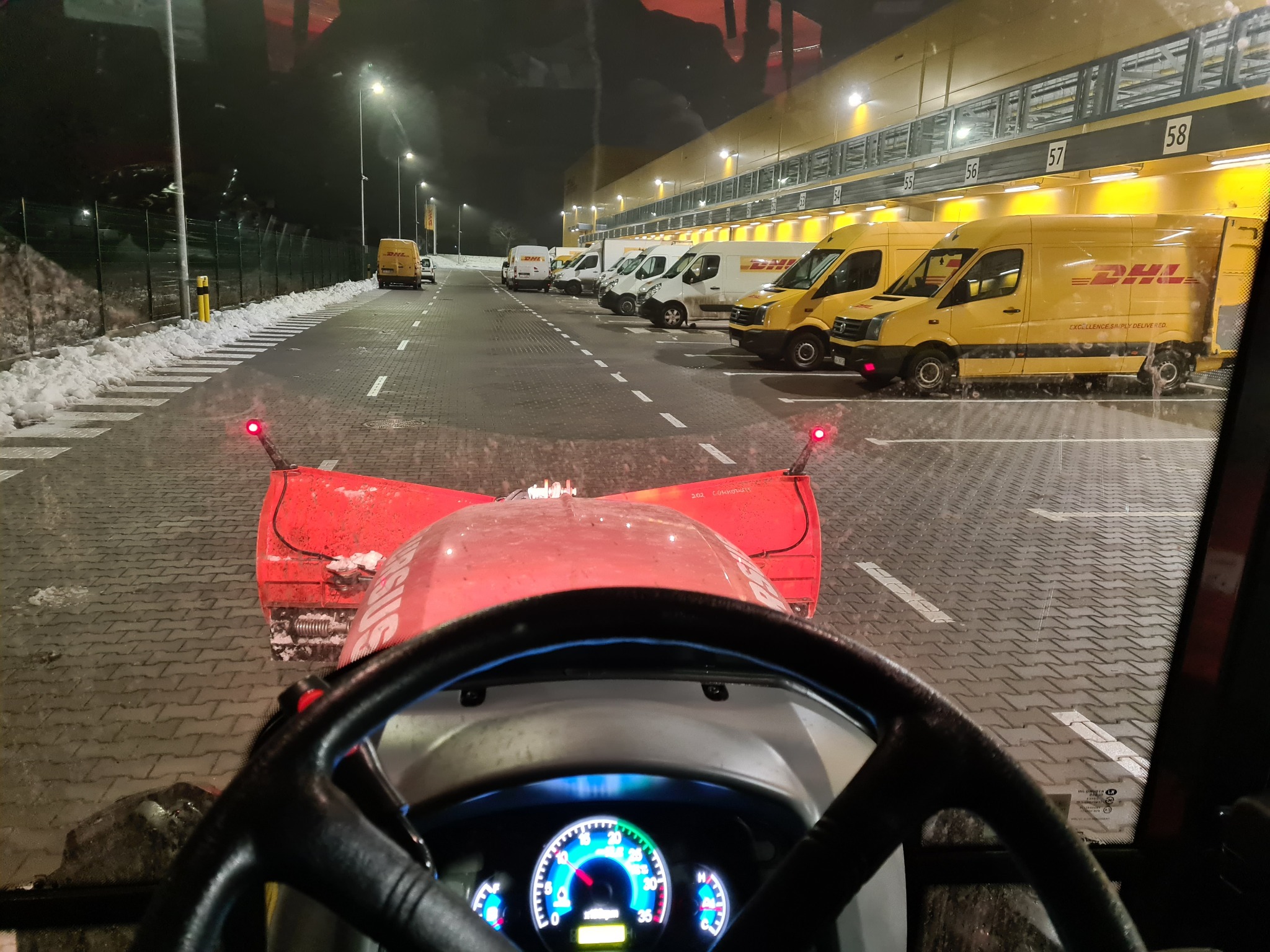 Widok z kabiny traktora z pługiem odśnieżającym, na placu z zaparkowanymi samochodami DHL w tle, nocą. Oświetlony teren, resztki śniegu przy krawędziach.