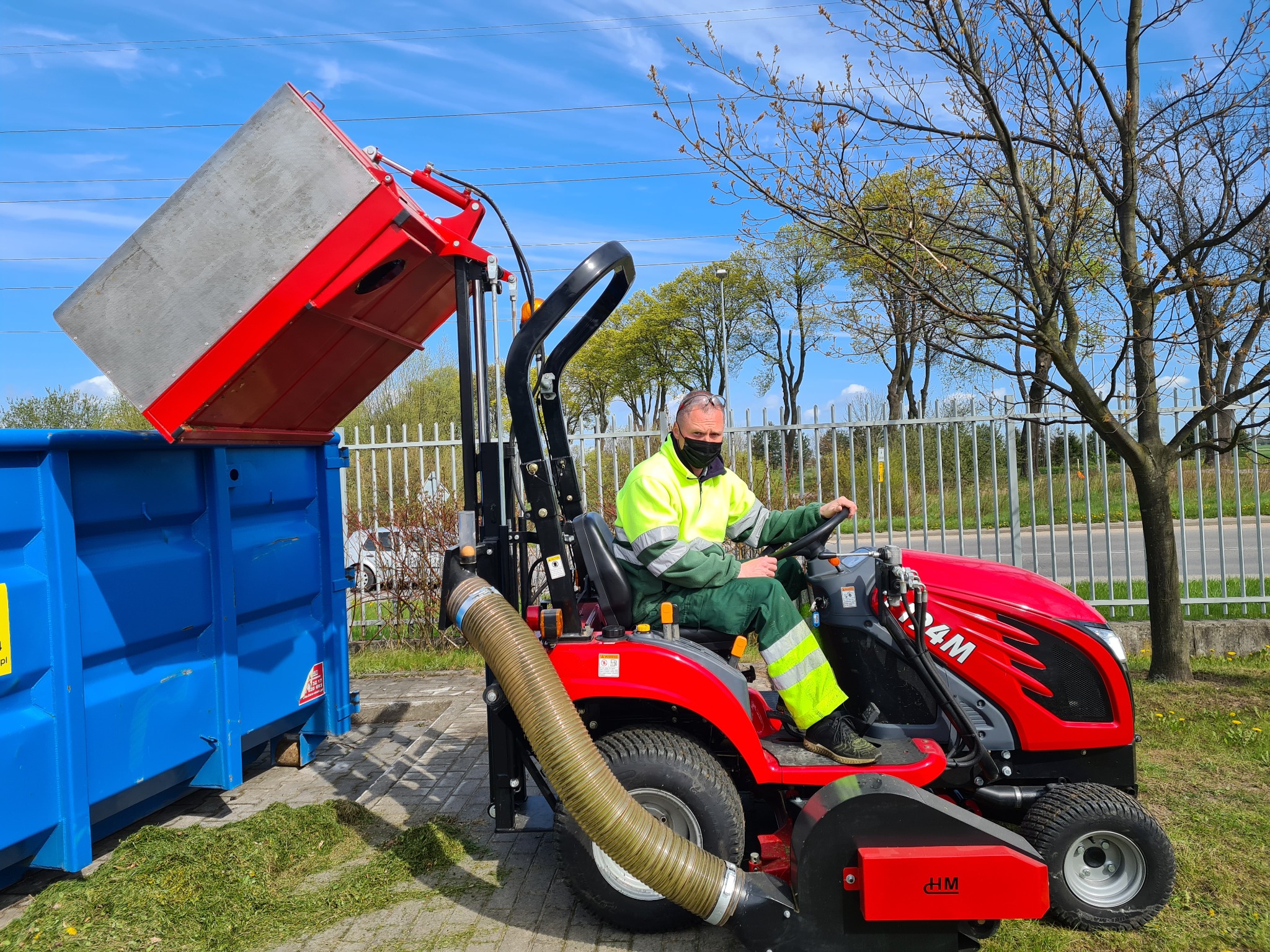 Operator w masce na czerwonym traktorze z odkurzaczem do trawy opróżnia zbiornik do niebieskiego kontenera na tle ogrodzenia i drzew.
