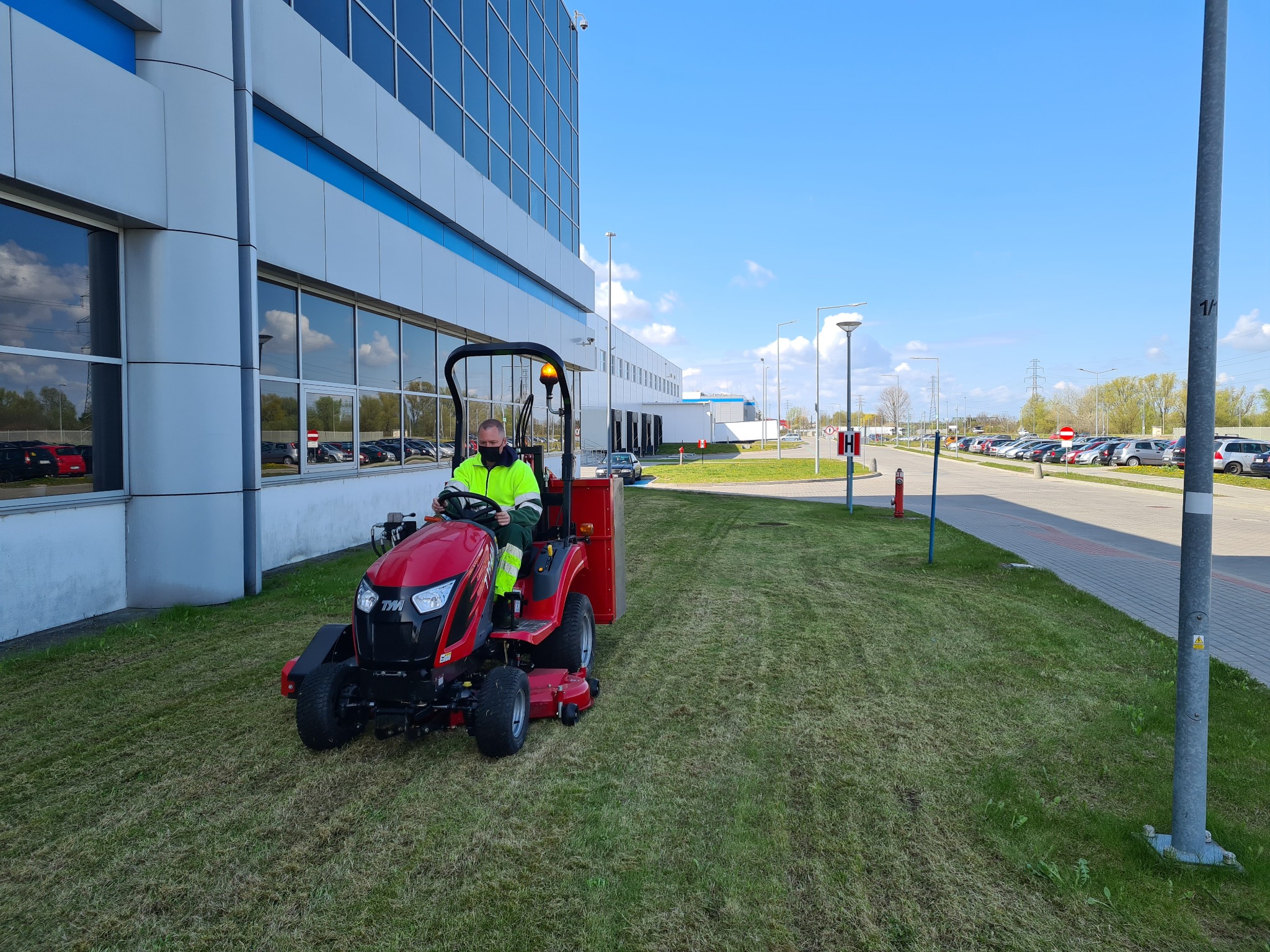 Czerwona kosiarka traktorek prowadzona przez operatora w odblaskowej kamizelce na tle nowoczesnego biurowca i parkingu. Widoczny efekt koszenia.