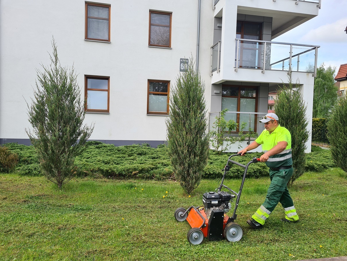 Mężczyzna w odblaskowej kamizelce kosiarką spalinową pielęgnuje trawnik przed nowoczesnym budynkiem z balkonem i ozdobnymi iglakami.