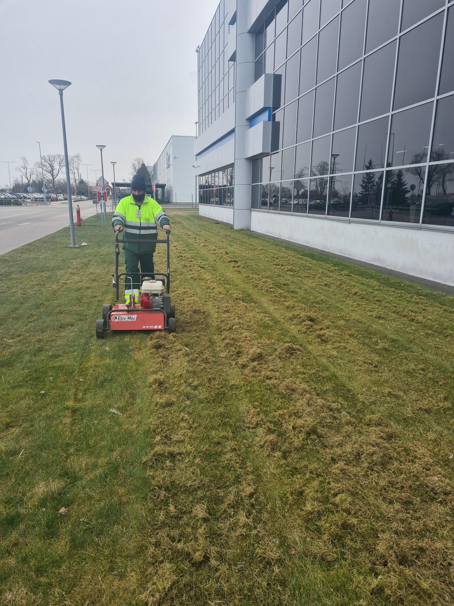 Wertykulacja trawnika przy budynku biurowym. Pracownik w odblaskowej kurtce obsługuje wertykulator OLEO-MAC na tle szklanej fasady.