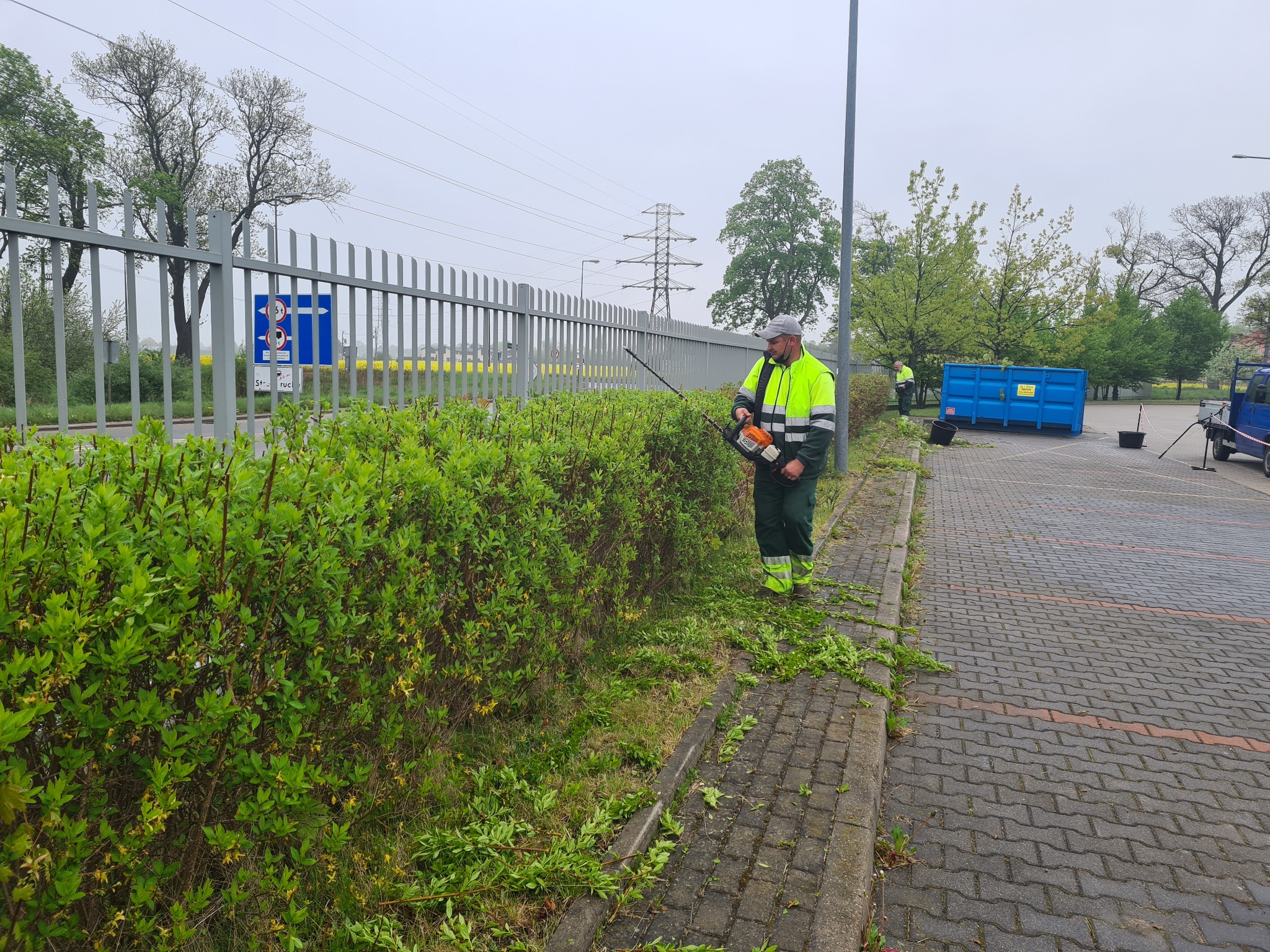Mężczyzna w odblaskowej kurtce przycina żywopłot przy użyciu nożyc spalinowych. W tle widoczny kontener i zaparkowany samochód dostawczy.