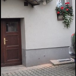 ClimaNext Gen Adam Jochemczyk - Biała jednostka klimatyzacji zamontowana na szarej ścianie budynku obok okna z kwiatami i brązowych drzwi wejściowych z daszkiem.