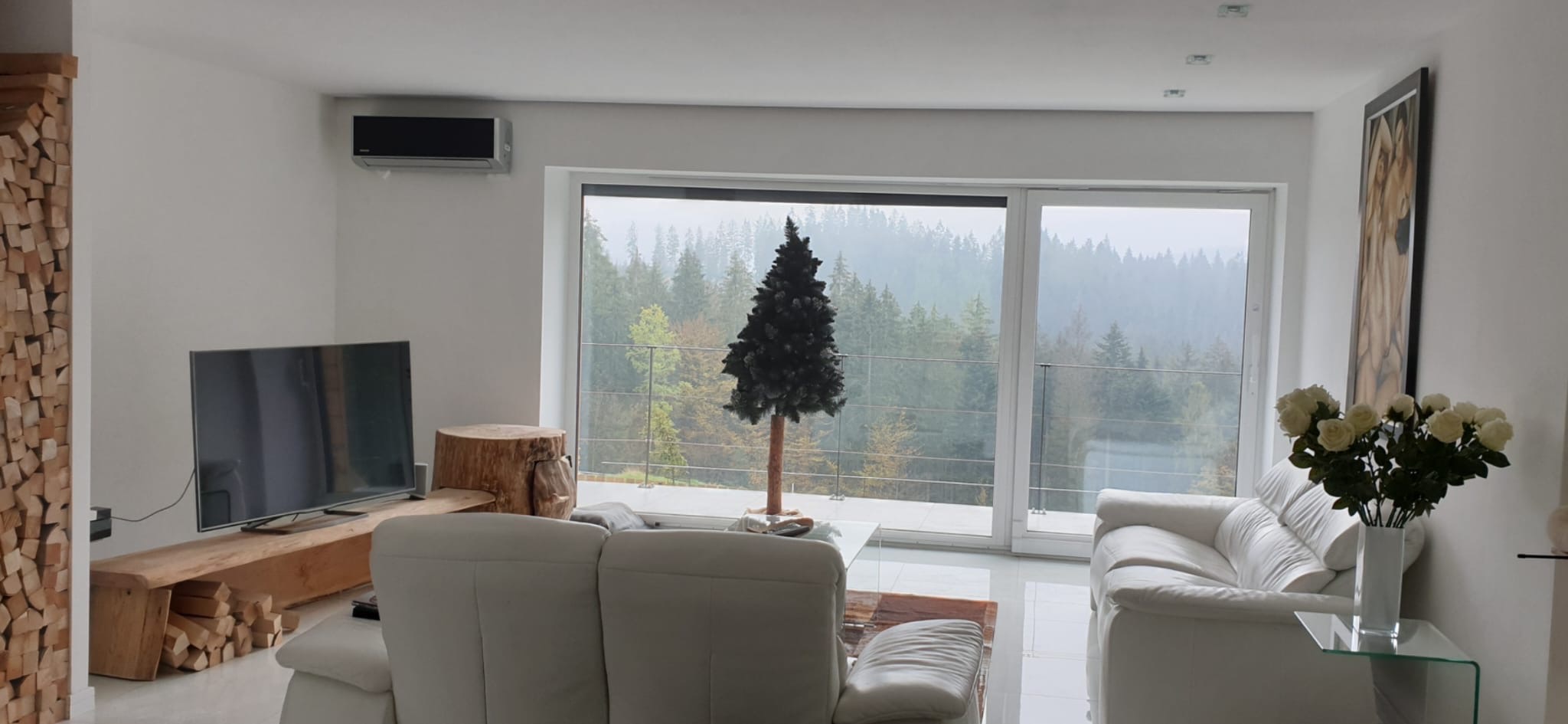 Nowoczesny salon z klimatyzatorem ściennym, widokiem na las i dekoracjami z drewna. Minimalistyczny design z białymi sofami i szklanym stolikiem.