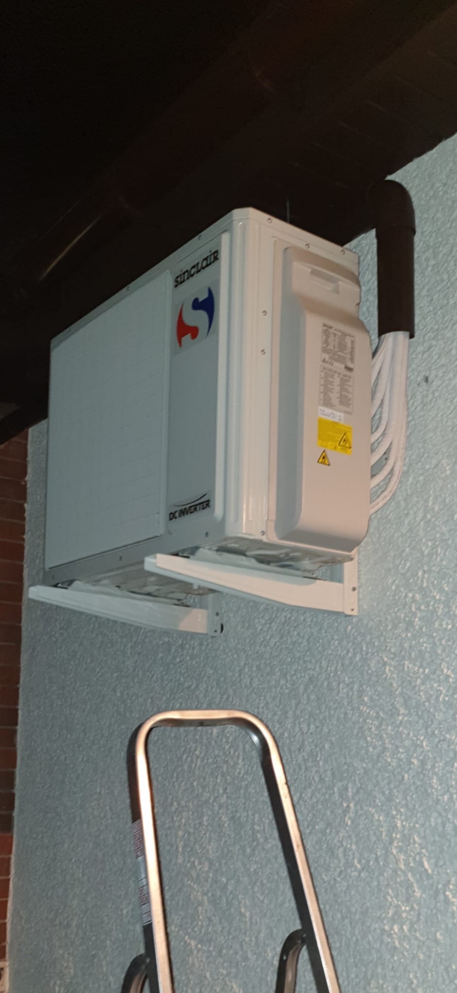 Zamontowana jednostka wewnętrzna klimatyzacji Sinclair DC Inverter na teksturowanej ścianie, widoczne rury instalacyjne i fragment drabiny.
