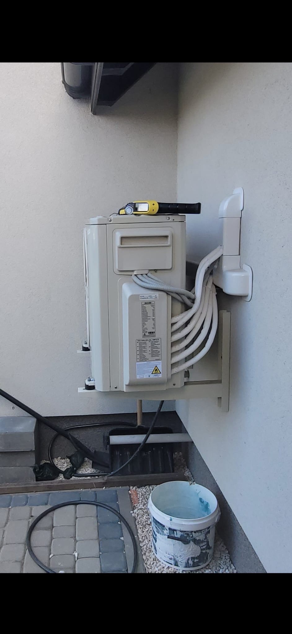 Zamontowana jednostka zewnętrzna klimatyzacji na ścianie budynku, z widocznymi przewodami i narzędziem pracy na górze. Widoczny fragment chodnika i wiadro.
