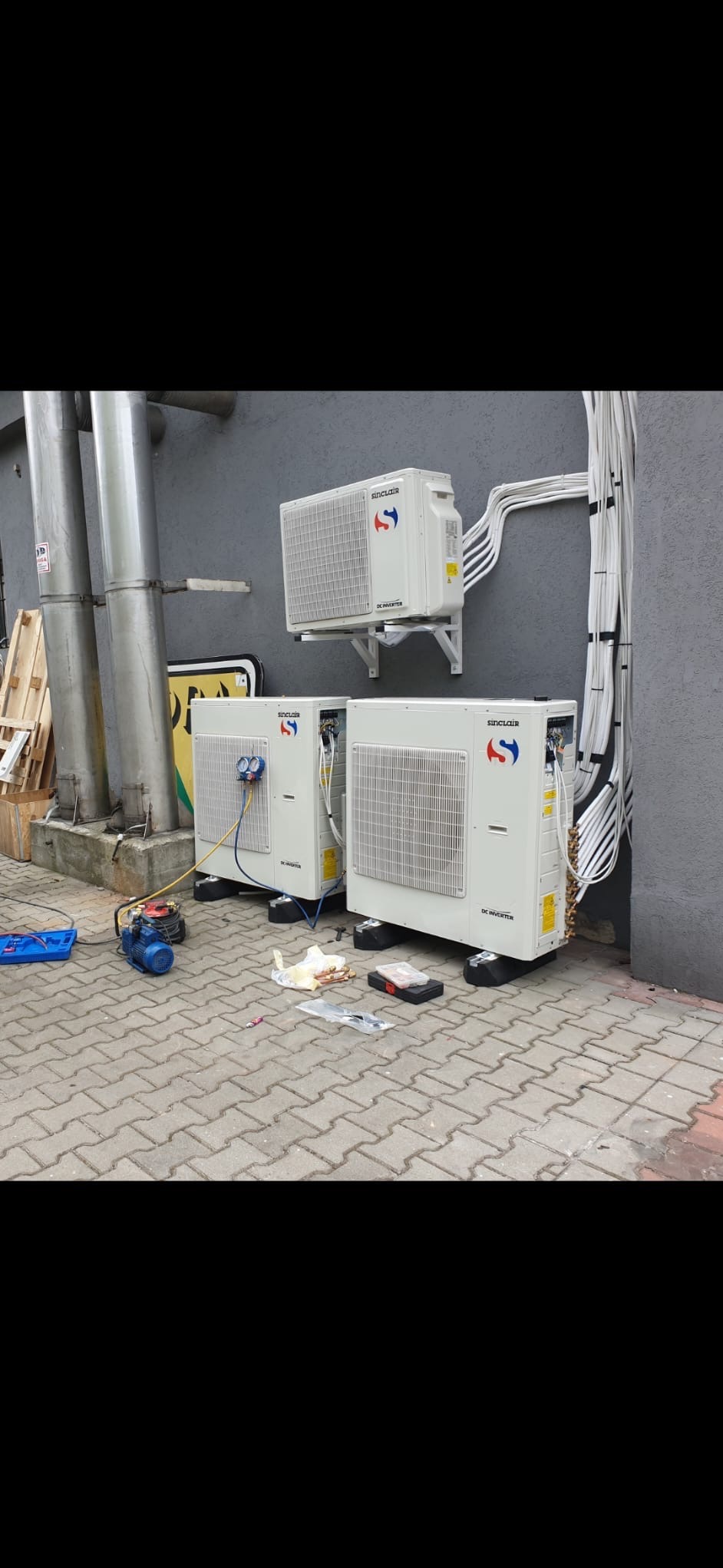 Montaż klimatyzacji Sinclair: trzy jednostki zewnętrzne, dwie stojące na kostce brukowej, jedna na wsporniku ściennym. Widoczne narzędzia i przewody.