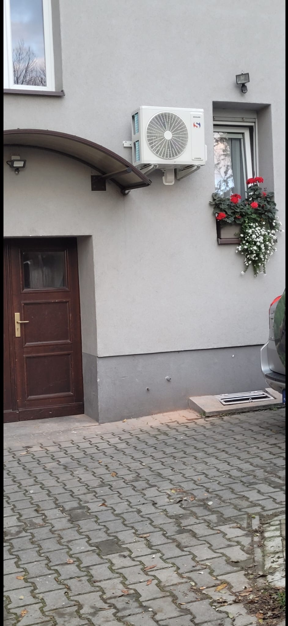 Biała jednostka klimatyzacji zamontowana na szarej ścianie budynku obok okna z kwiatami i brązowych drzwi wejściowych z daszkiem.