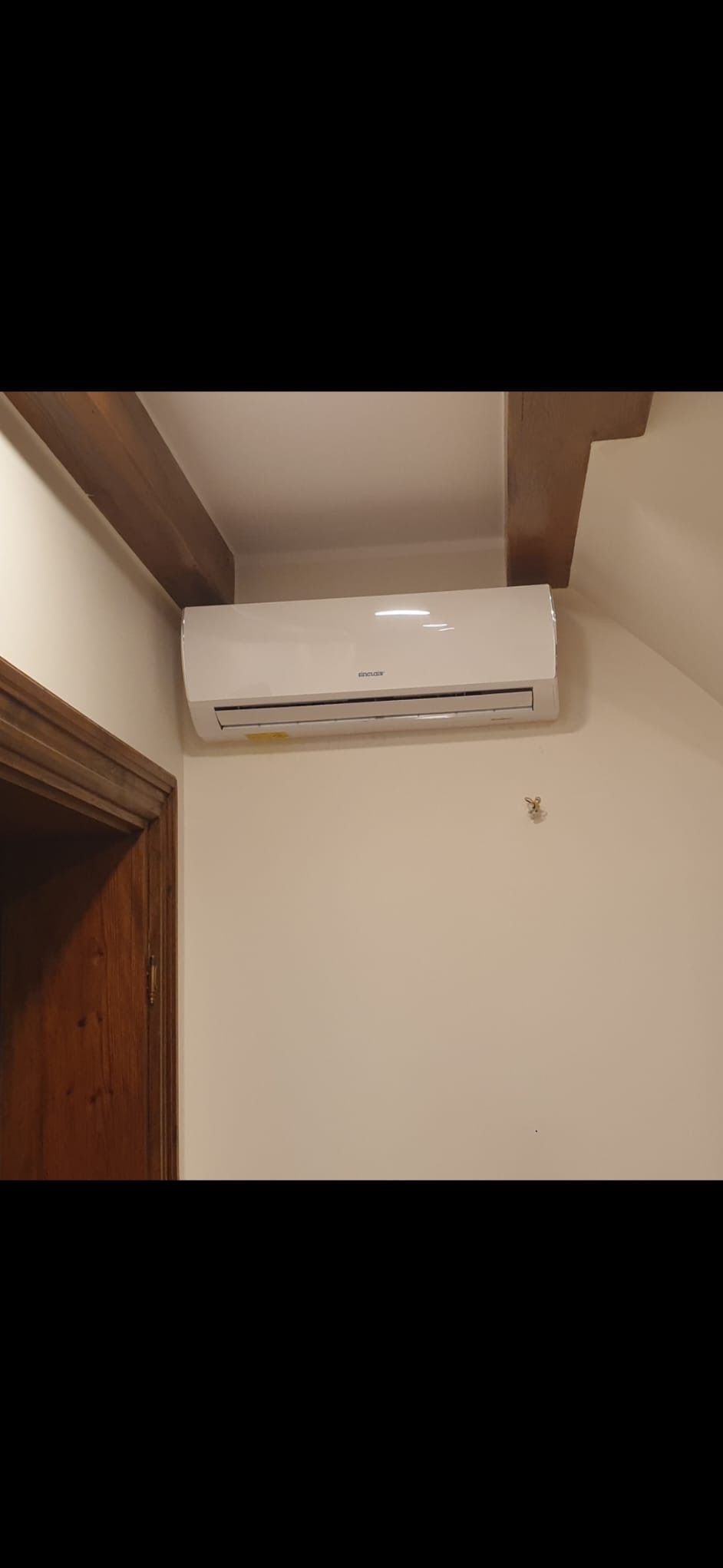 Biały klimatyzator Sinclair zamontowany na ścianie w pomieszczeniu z drewnianymi belkami stropowymi i drzwiami. Widok z dołu.
