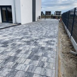 Świat-Bruk - Ułożona kostka brukowa w odcieniach szarości przed nowoczesnym domem z czarną stolarką okienną, widok z perspektywy chodnika.