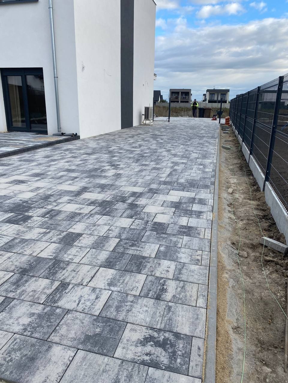 Ułożona kostka brukowa w odcieniach szarości przed nowoczesnym domem z czarną stolarką okienną, widok z perspektywy chodnika.