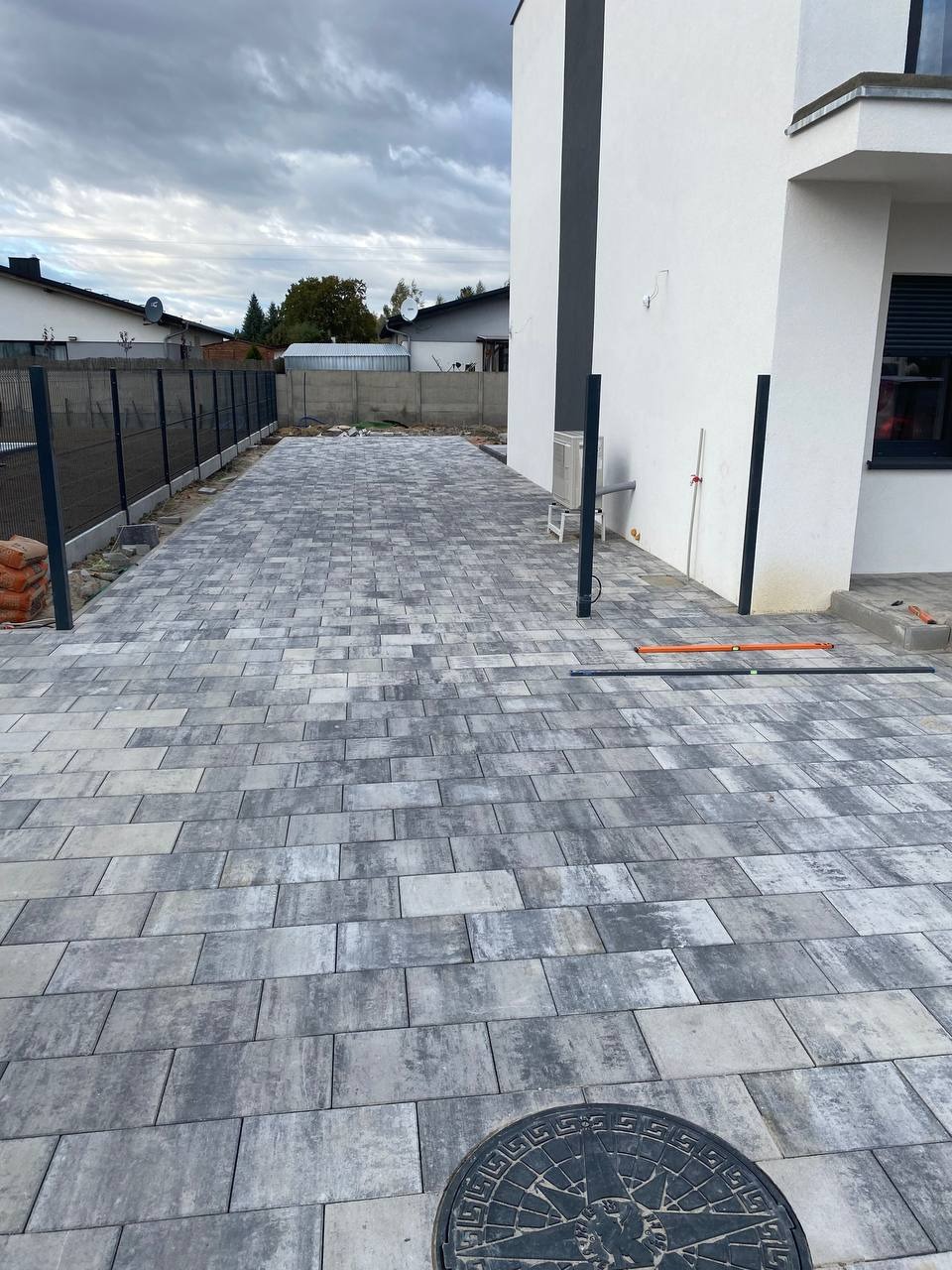 Ułożona szara kostka brukowa na podjeździe domu z nowoczesną elewacją, widoczne narzędzia i studzienka kanalizacyjna. Całość w pochmurny dzień.