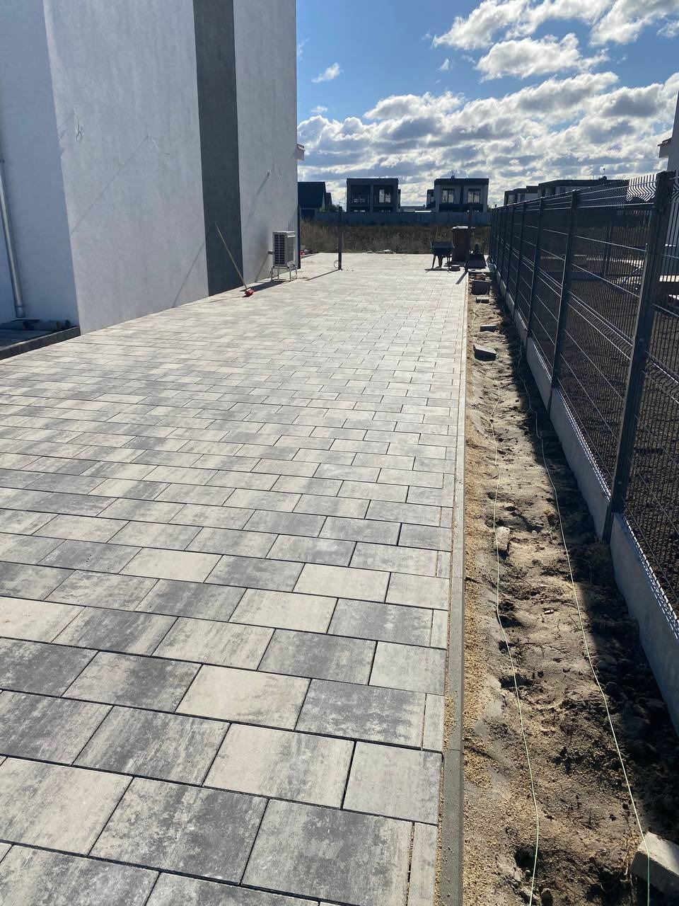 Ułożona, szara kostka brukowa na posesji z metalowym ogrodzeniem i budynkiem w tle, widok z perspektywy.