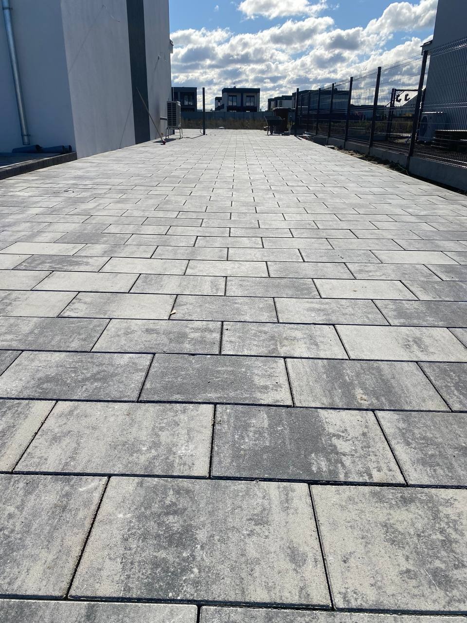 Ułożona kostka brukowa w odcieniach szarości tworząca długi chodnik wzdłuż budynku z metalowym ogrodzeniem. Widok z perspektywy chodnika.