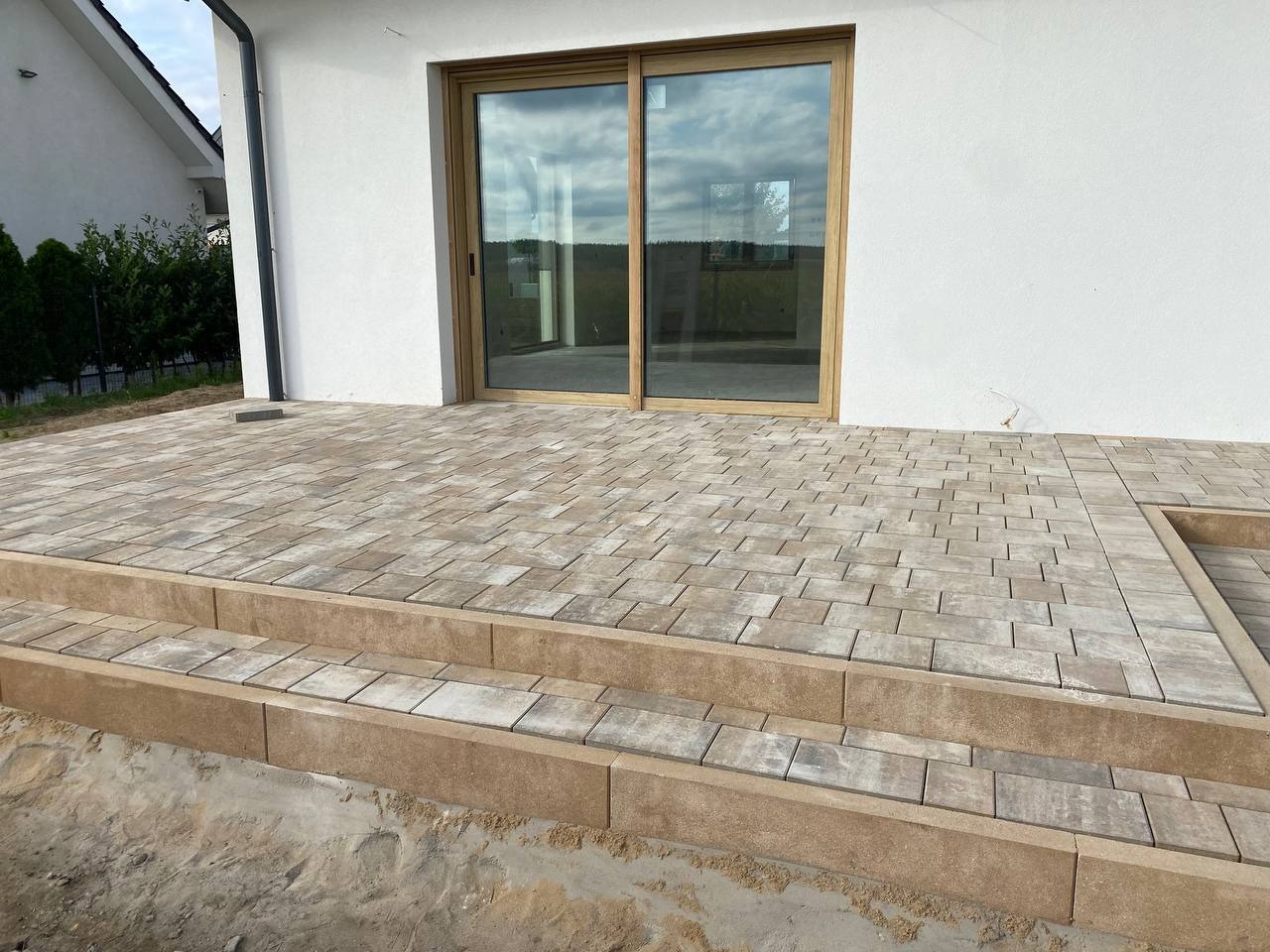 Wykończony taras z kostki brukowej w odcieniach beżu i szarości, z dwustopniowymi schodami przy białej ścianie domu z przesuwnymi drzwiami tarasowymi.