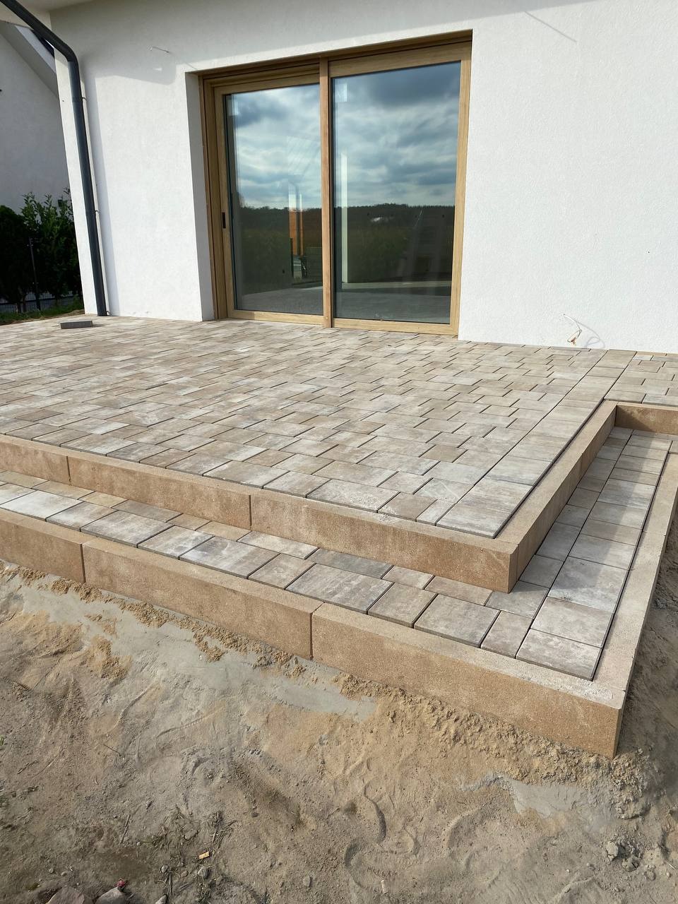 Wyłożony kostką taras z dwoma stopniami przy białym budynku z dużym oknem przesuwnym. Widoczny piasek wokół, co sugeruje świeżo zakończone prace.