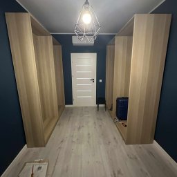 Bartosz Dąbek - Wnętrze z dwoma jasnymi, drewnianymi regałami po bokach, niebieską ścianą i wiszącą lampą. Na podłodze leżą narzędzia i plany, sugerując trwający montaż.