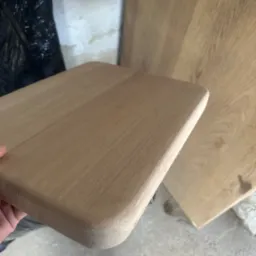 Drewniany blat z zaokrąglonymi krawędziami, ukazujący strukturę drewna. W tle widoczny fragment większej płyty drewnianej i czarny worek.