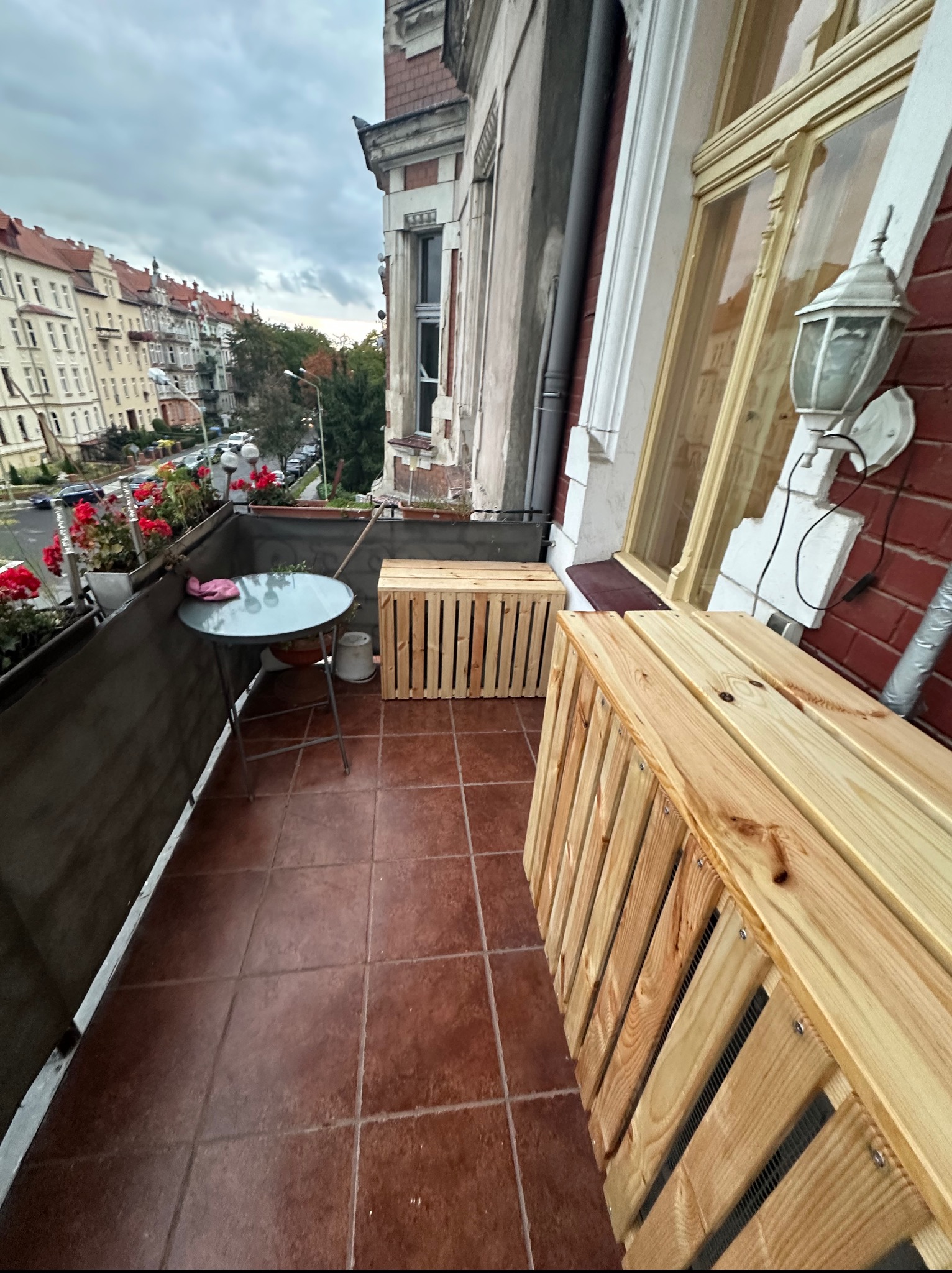 Umeblowany balkon z drewnianymi skrzyniami, stolikiem i kwiatami w donicach, z widokiem na ulicę i budynki w tle. Czerwona cegła i lampa na ścianie.