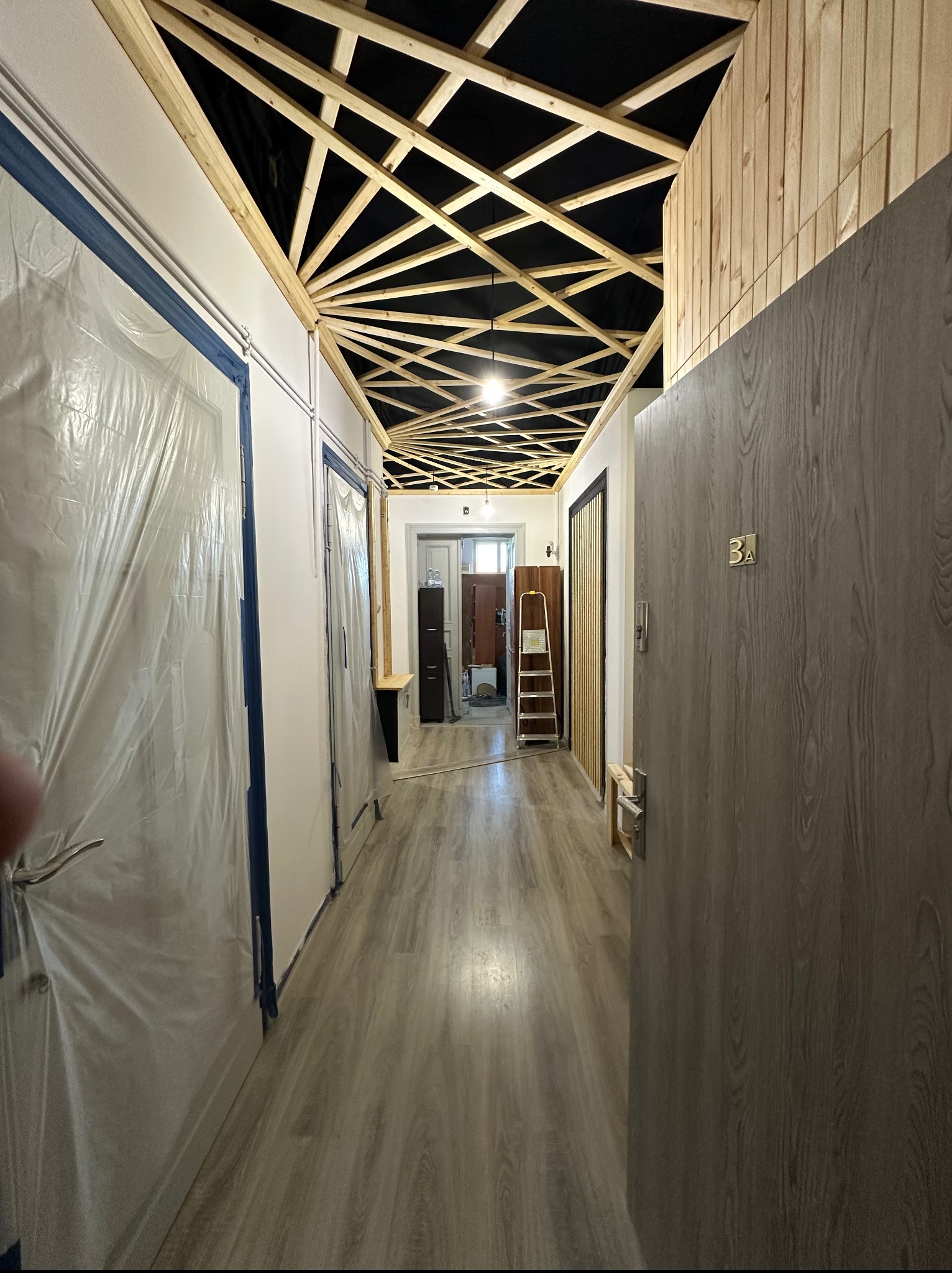 Korytarz w trakcie remontu z drewnianą konstrukcją sufitu, zabezpieczone folią drzwi, drewniane panele ścienne i podłogowe. Widoczna drabina i otwarte drzwi.