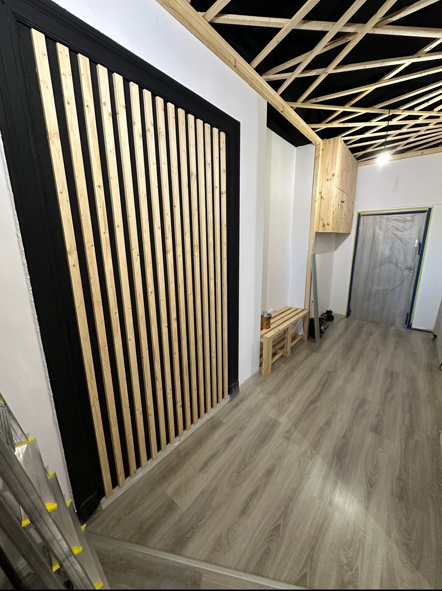 Nowoczesna aranżacja wnętrza z drewnianymi lamelami na ścianie w czarnej ramie, widoczny sufit z drewnianą konstrukcją i jasna podłoga. Częściowo widoczna drabina.