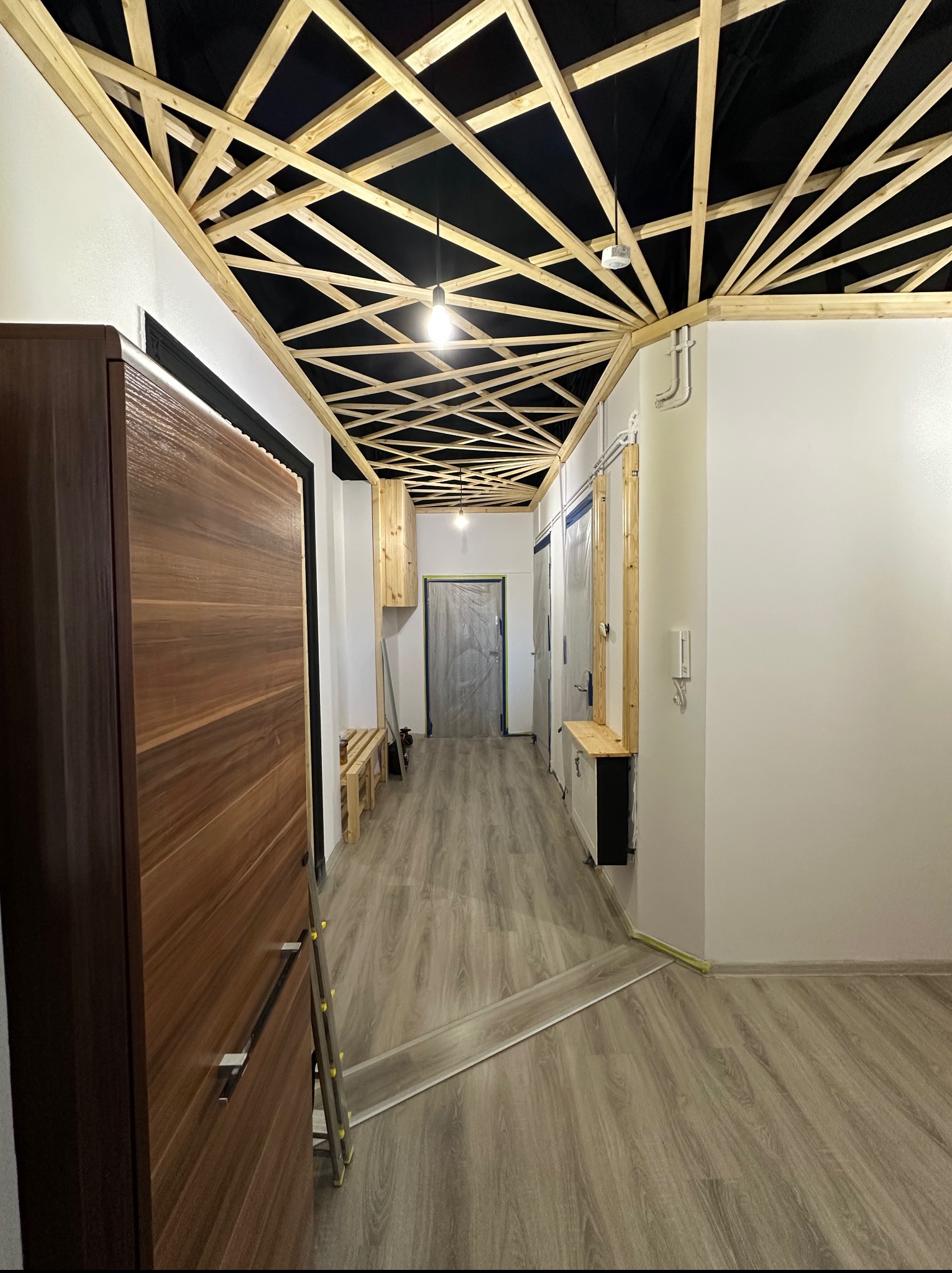 Wykończenie sufitu z drewnianą konstrukcją i oświetleniem w korytarzu. Widoczne drzwi, podłoga z paneli i fragment ściany. Użyte materiały i oświetlenie.
