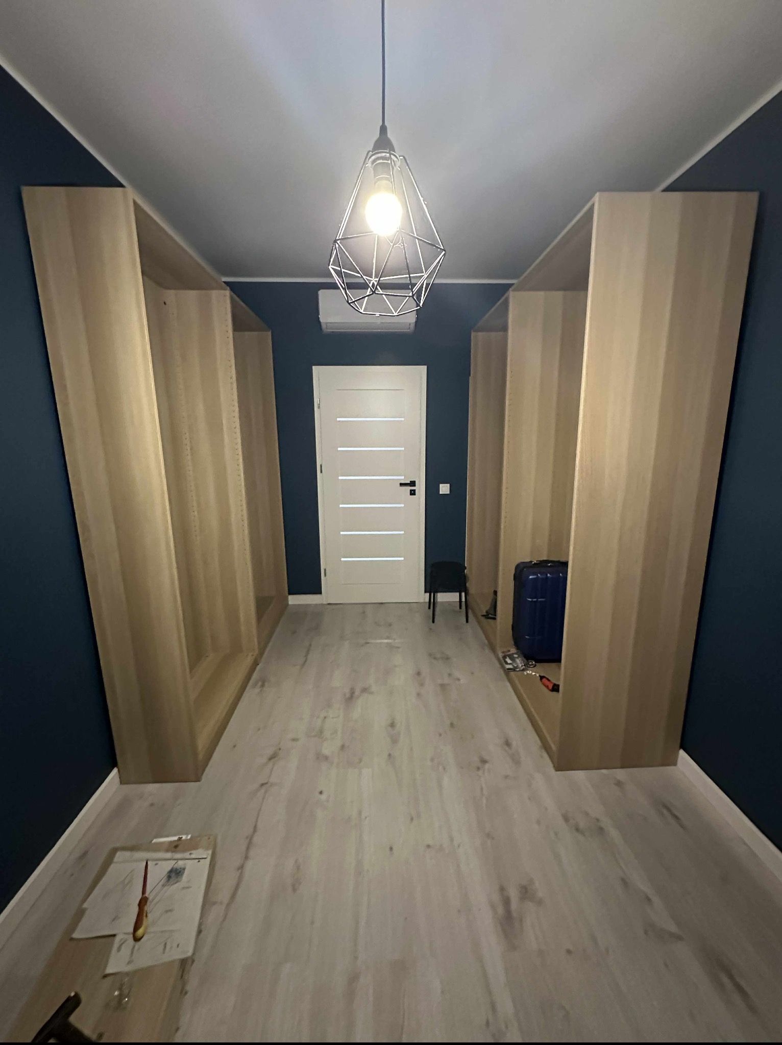 Wnętrze z dwoma jasnymi, drewnianymi regałami po bokach, niebieską ścianą i wiszącą lampą. Na podłodze leżą narzędzia i plany, sugerując trwający montaż.