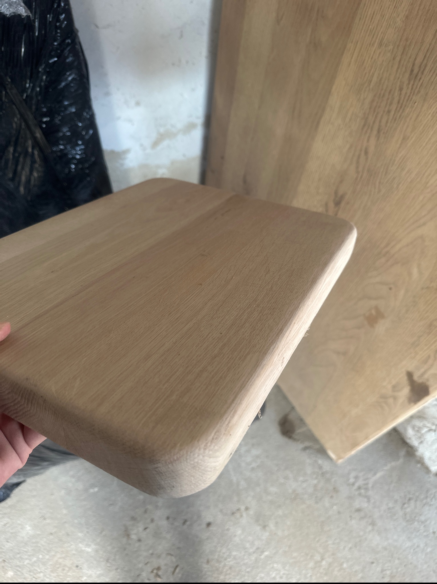 Drewniany blat z zaokrąglonymi krawędziami, ukazujący strukturę drewna. W tle widoczny fragment większej płyty drewnianej i czarny worek.
