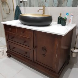 Renowacja Mebli Dominik Zaskórski - Stylowa, ciemnobrązowa szafka łazienkowa z marmurowym blatem i owalną umywalką w kolorze czarno-złotym. Widoczna armatura i akcesoria łazienkowe.