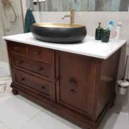 Stylowa, ciemnobrązowa szafka łazienkowa z marmurowym blatem i owalną umywalką w kolorze czarno-złotym. Widoczna armatura i akcesoria łazienkowe.