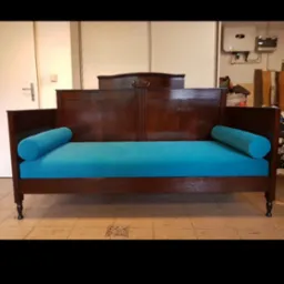 Antyczny, odnowiony tapczan z ciemnego drewna i turkusową tapicerką. Widoczne charakterystyczne nogi i dekoracyjne elementy rzeźbione. Tło: ściana i fragment instalacji.