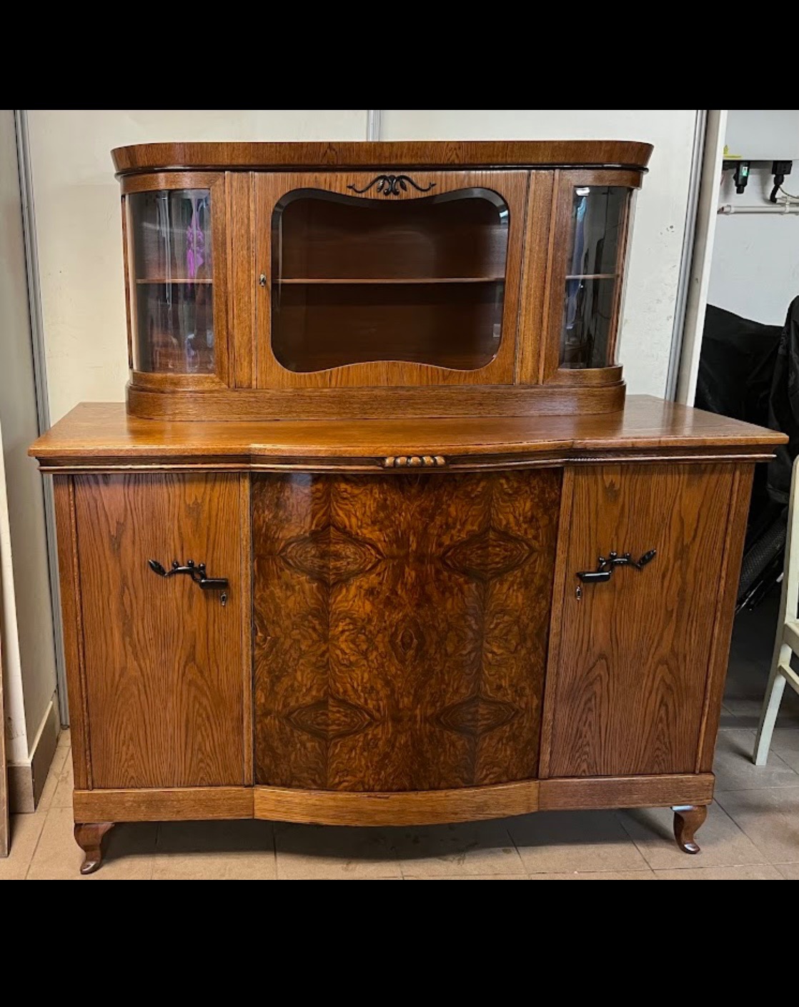 Antyczna, fornirowana komoda z nadstawką i szklanymi drzwiczkami, z dekoracyjnymi uchwytami, w ciepłych odcieniach brązu i beżu, na niskich nóżkach.