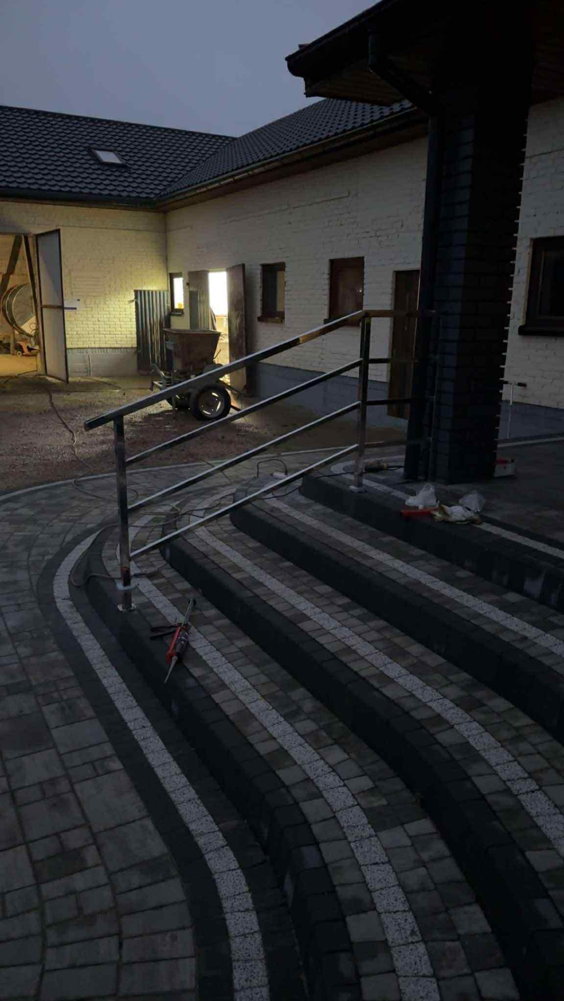 Nierdzewna balustrada schodowa z trzema poziomami, zamontowana na zewnątrz budynku z kostki brukowej. Widoczne narzędzia montażowe.