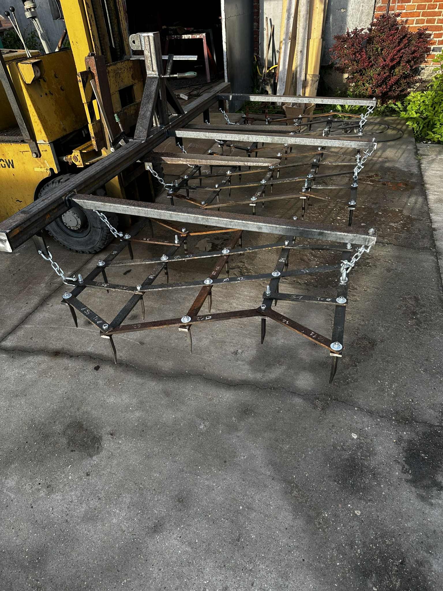 Metalowa brona z łańcuchami, podwieszona na żółtym wózku widłowym, na betonowym podłożu. Widoczne ślady spawania i montażu. Tło stanowi ceglany budynek i zieleń.