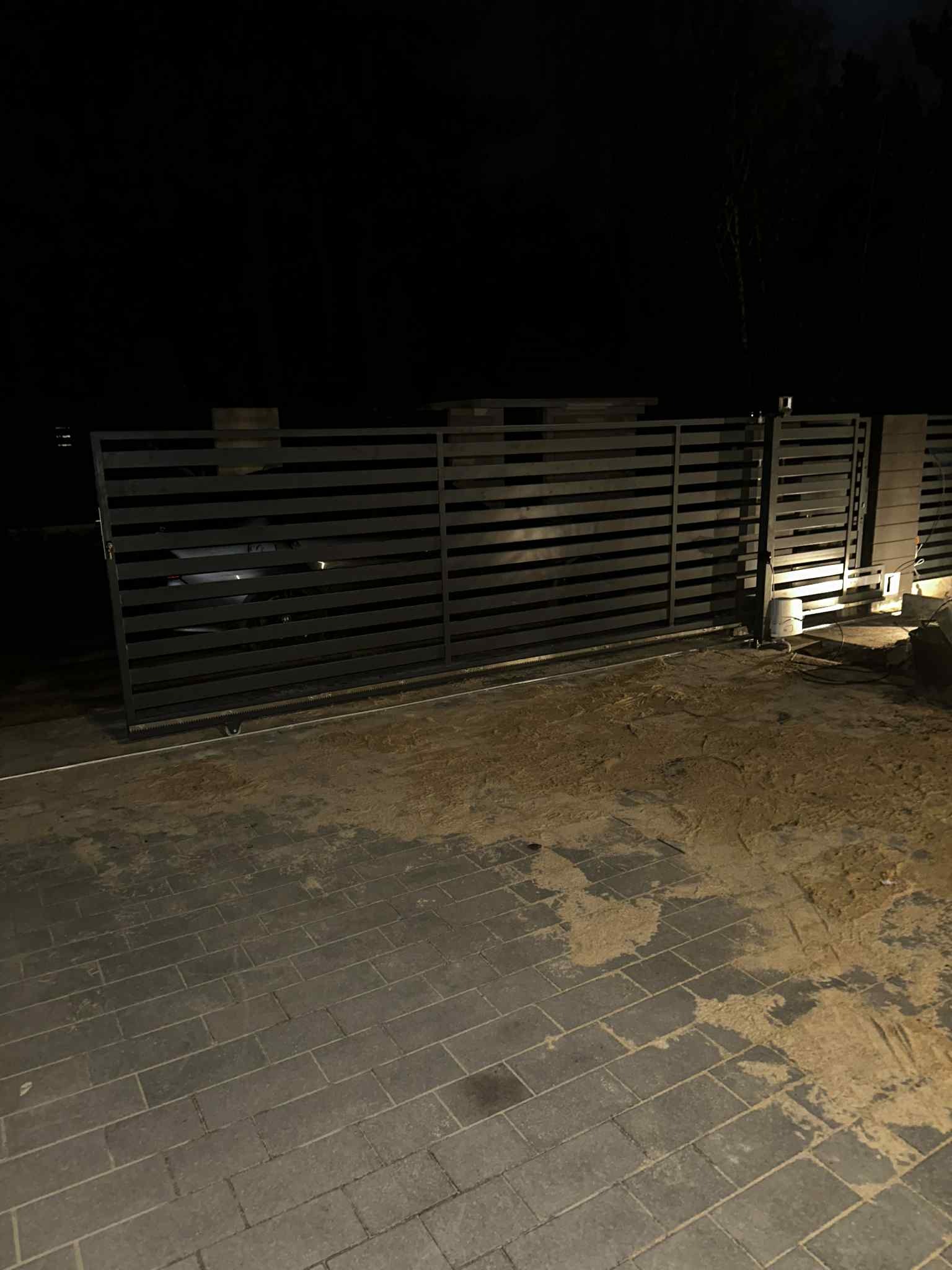 Nowoczesna, przesuwna brama wjazdowa z poziomymi panelami, oświetlona nocą. Widoczny mechanizm automatycznego otwierania i fragment podjazdu z kostki brukowej.