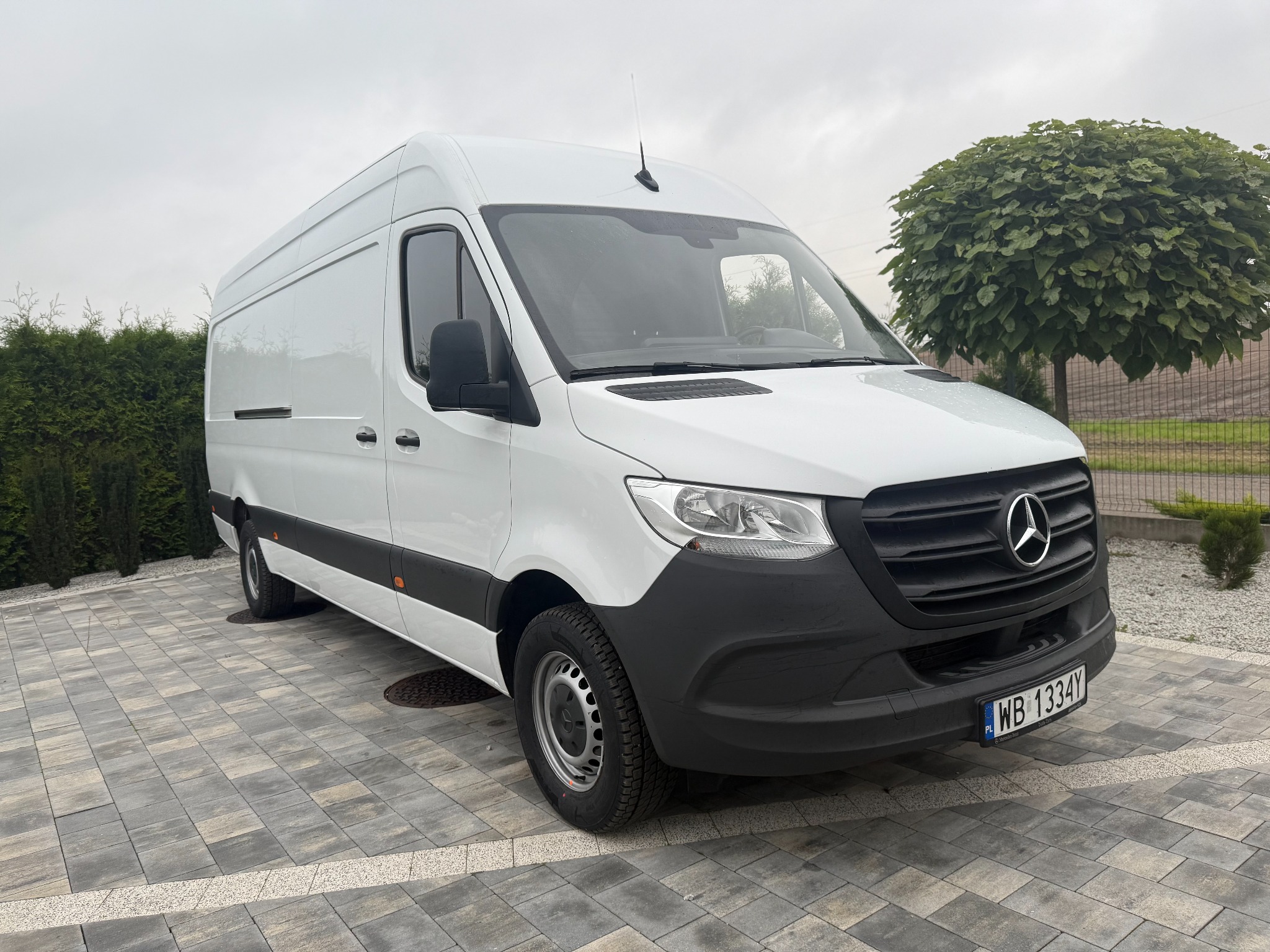 Biały samochód dostawczy Mercedes Sprinter zaparkowany na szarej kostce brukowej, z zielonymi krzewami w tle i pochmurnym niebem. Widoczny numer rejestracyjny.